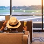 man traveler with hat holding beer glass and looking to airplane, Asian passenger sitting and relax in modern lounge at international airport terminal. Travel concept