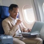 Elegant businessman using laptop while sitting in airplane cabin with sun shining trough airplane window.