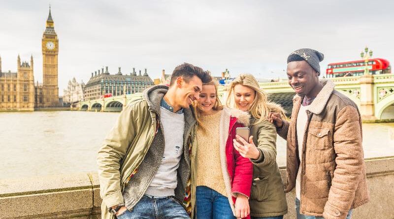 Happy multiracial friends group using smartphone in London. Mixed race millennials people lifestyle concept. Friends sharing trip on social network. Big ben and Westminster parliament on background.