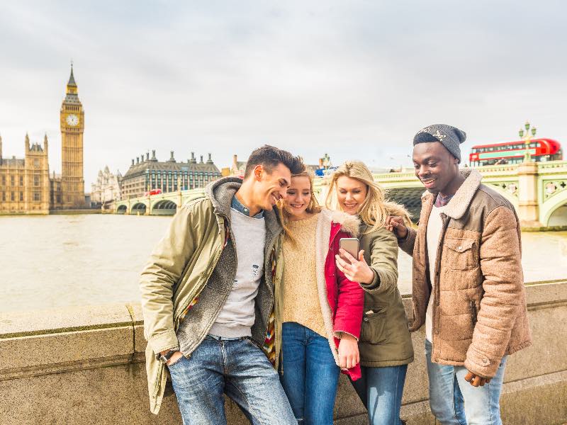 Happy multiracial friends group using smartphone in London. Mixed race millennials people lifestyle concept. Friends sharing trip on social network. Big ben and Westminster parliament on background.