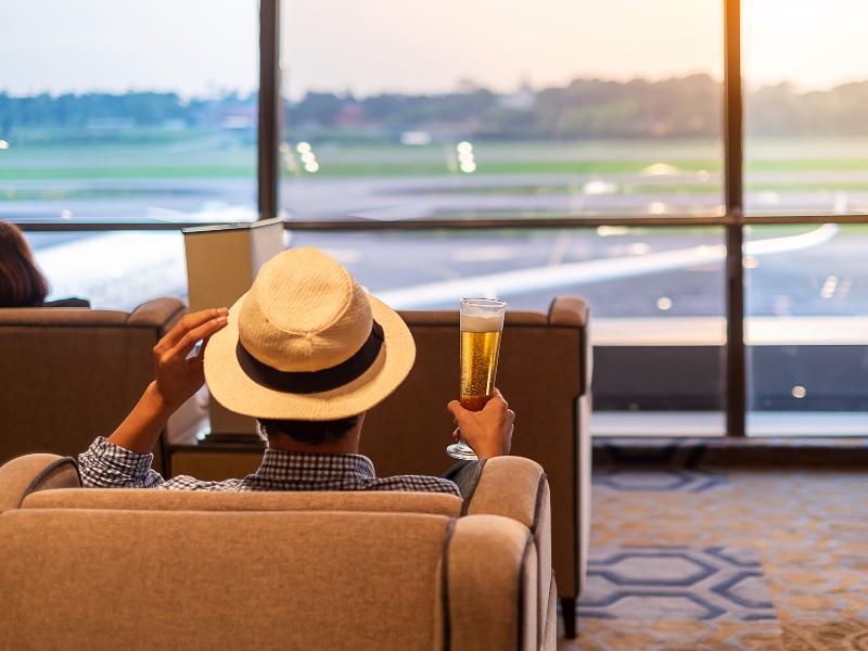 man traveler with hat holding beer glass and looking to airplane, Asian passenger sitting and relax in modern lounge at international airport terminal. Travel concept